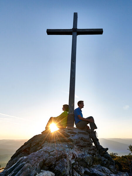 Paar sitzt am Gipfelkreuz eines Berges bei Sonnenschein