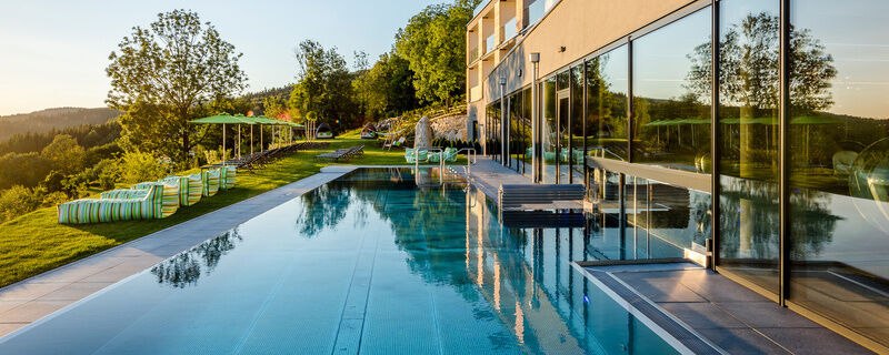 Außenansicht Hotel mit Außenpool, Liegen und Sonnenschirme auf Wiese mit Wald im Hintergrund