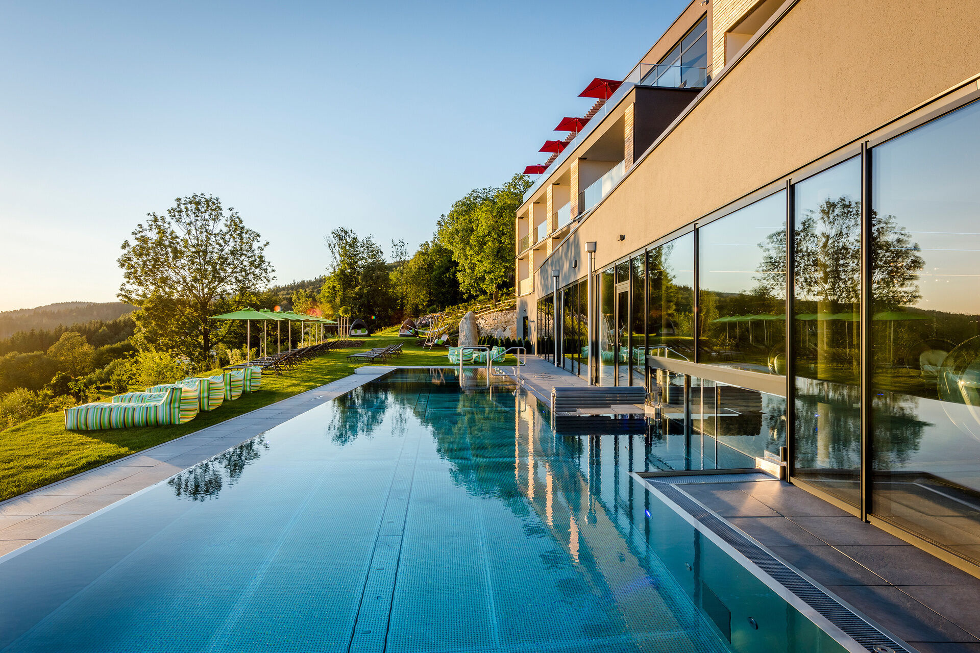 Außenansicht Hotel mit Außenpool, Liegen und Sonnenschirme auf Wiese mit Wald im Hintergrund