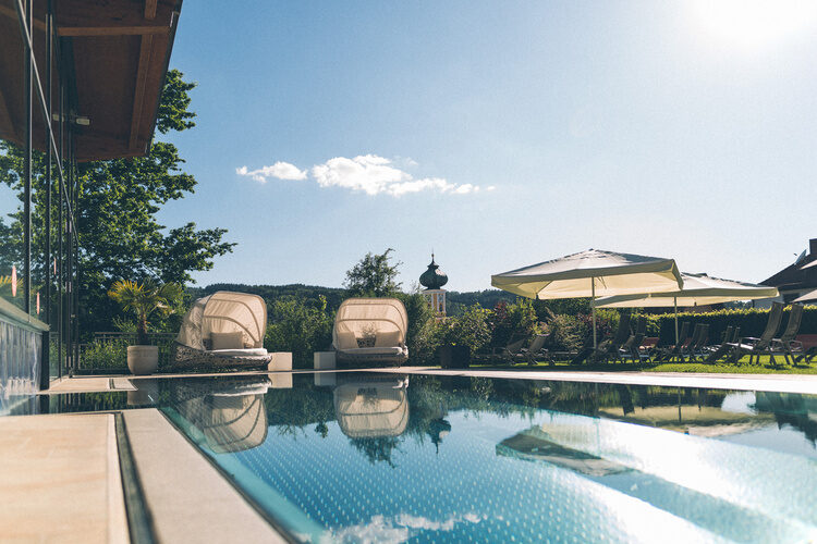 Spiegelnder Außen-Pool bei warmen Sonnenschein, im Hintergrund gemütliche Sonnenliegen mit Sonnenschirm im Hotel Garten.