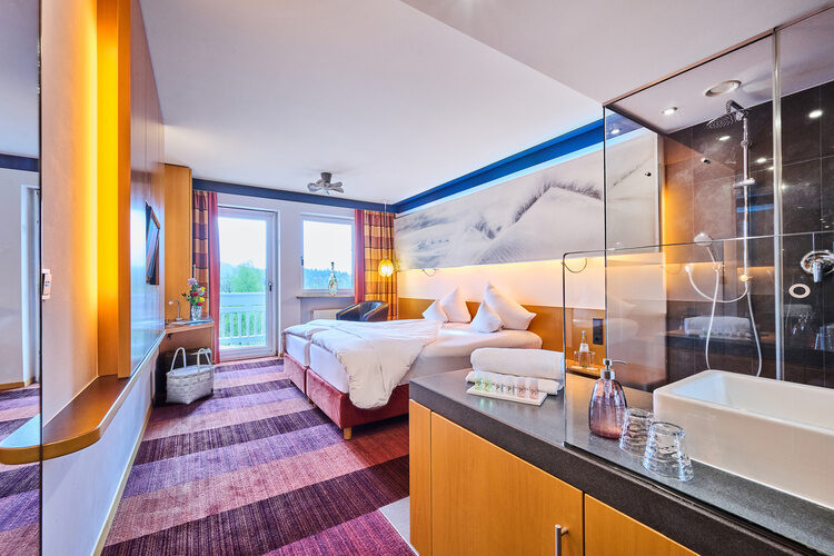 Hotelzimmer mit Doppelbett, gläserner Walk-in-Dusche, Waschbecken und Balkonfenster.
