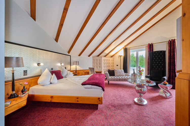 Geräumige Suite mit Dachschrägen, rotem Teppich, Doppelbett, Sitzmöbeln und Balkontür.