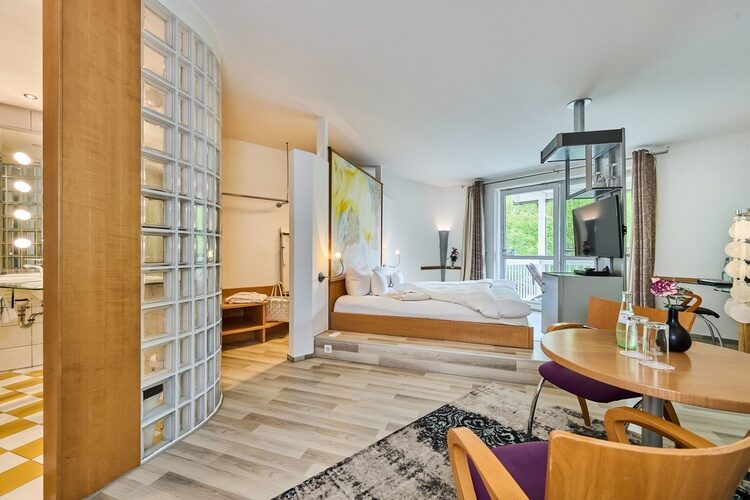 Moderne Dachzimmer-Suite mit Glasblock-Badwand, erhöhtem Bett, Esstisch, Stuhl und Balkon.