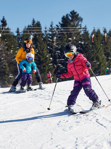 Mädchen beim Skifahren mit Frau und Kleinkind im Hintergrund beim Lernen des Skifahrens